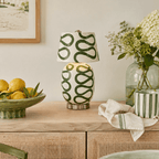 The green-looped version of this Clérachic table lamp stands on a cane-fronted sideboard beside a lemon bowl, a glass vase and white flowers.