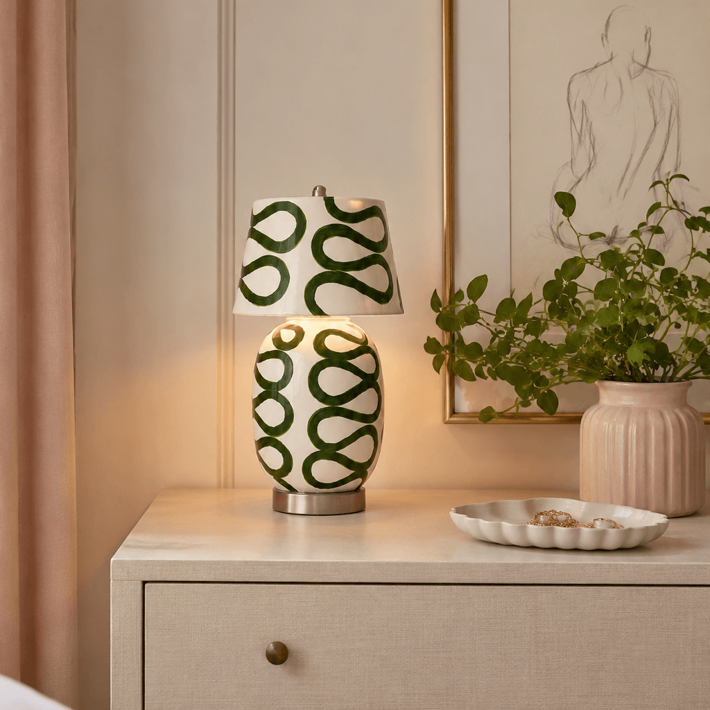 This Clérachic table lamp in cream and green glows softly on a white chest of drawers beside a pink fluted vase and a line drawing on the wall.