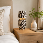 Black-and-white zebra-stripe ceramic body and shade on this Clérachic table lamp, lit warm on a wooden bedside table next to a bed and a white vase.