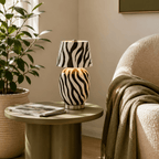 A striped black-and-white Clérachic table lamp stands on a round olive-green side table with a magazine, beside a boucle armchair and a leafy plant.