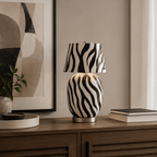 A zebra-striped Clérachic table lamp sits on a dark wooden sideboard in front of a large abstract print, with a stack of books to the side.