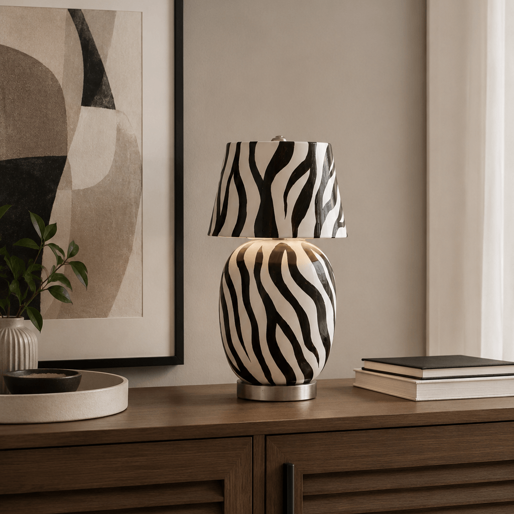 A zebra-striped Clérachic table lamp sits on a dark wooden sideboard in front of a large abstract print, with a stack of books to the side.