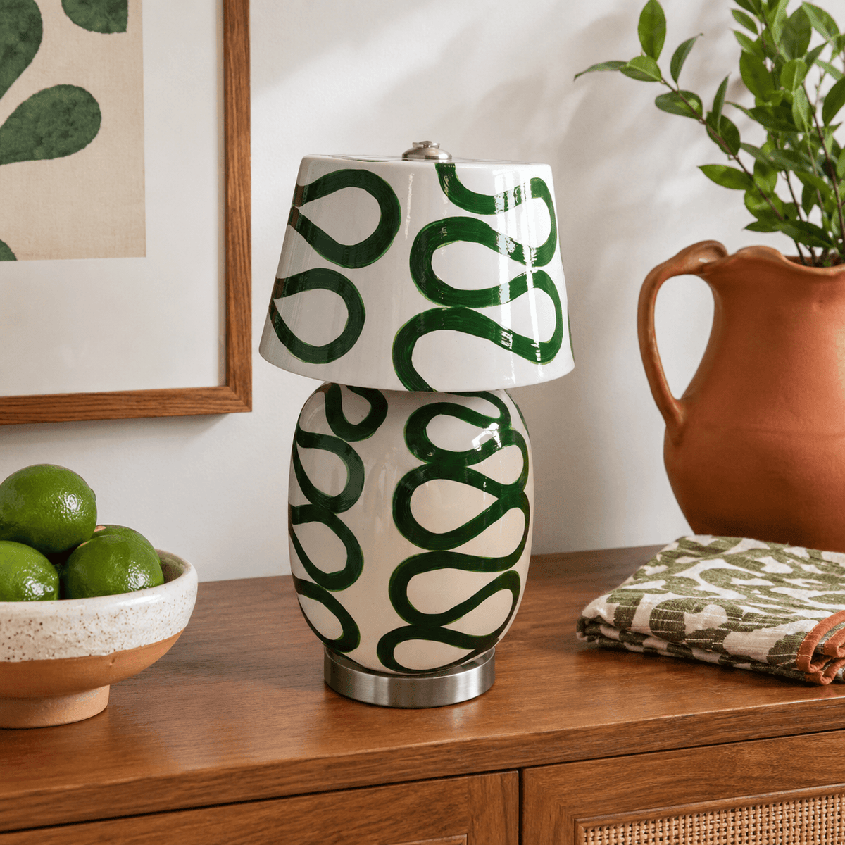 Close view of the glossy green loops painted on the body and shade of this Clérachic table lamp, on a wooden sideboard beside a terracotta jug and limes.