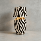 The striped version of this Clérachic table lamp stands on a pale concrete surface, its body glowing faintly through the black-and-white pattern.