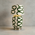 The cream and dark-green version of this Clérachic table lamp, lit, on a pale concrete surface in front of a grey plaster wall.