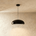 Clérachic glossy black dome pendant on a slim black cord, lit from beneath against a limewashed plaster wall.
