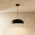 Clérachic glossy black dome pendant on a slim black cord, lit from beneath against a limewashed plaster wall.