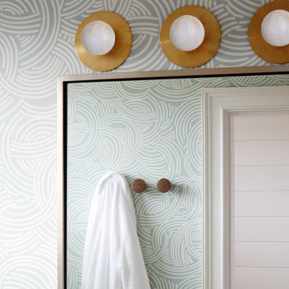 Three Clérachic Glow sconces in brass above a mirror on patterned green-and-white wallpaper.