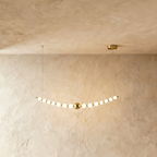 Clérachic linear chandelier, a gently curved gold-finished rod of opal spherical shades with a brass ball at the centre, lit.