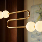 Close view of the Clérachic S-shaped linear chandelier, a polished gold-finished tube bend between glowing opal spherical shades.