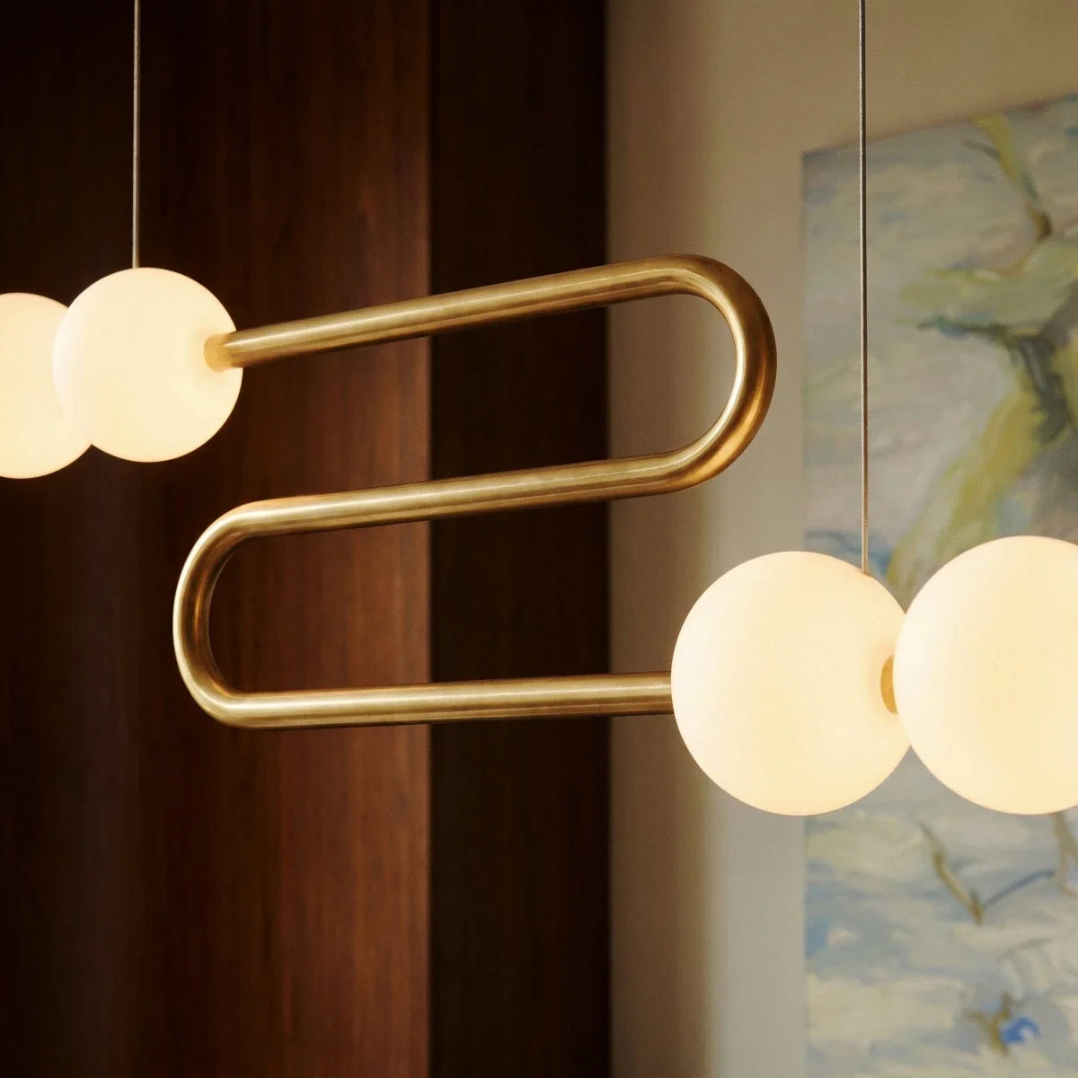Close view of the Clérachic S-shaped linear chandelier, a polished gold-finished tube bend between glowing opal spherical shades.