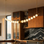 Clérachic curved linear chandelier close up, lit opal globes along a gold-finished rod with a central brass ball in a kitchen.