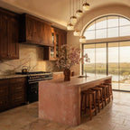 Six Clérachic alabaster pendant lights cascade above a stone kitchen island beneath an arched window at sunset.
