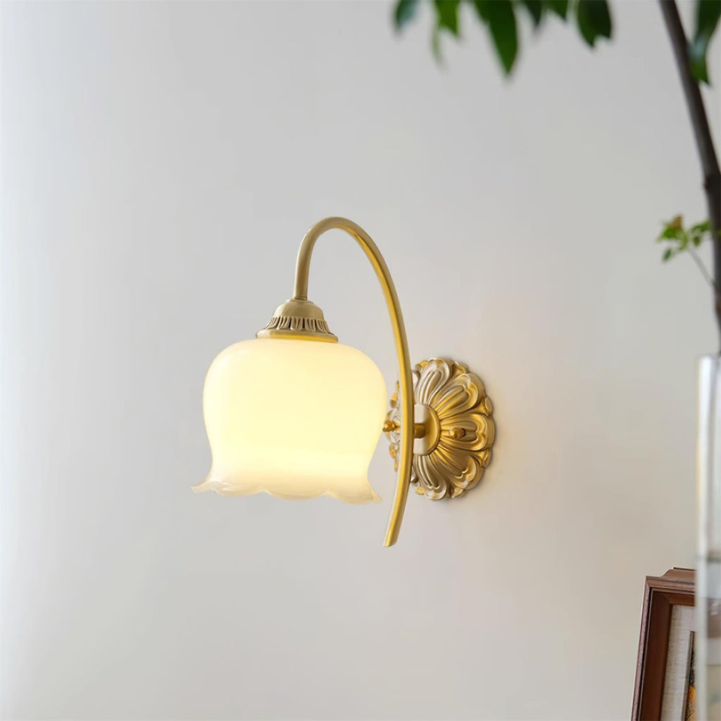 Unlit cream glass flower shade on a curved brass arm, a Clérachic wall light on a pale wall with a plant leaf at the edge.