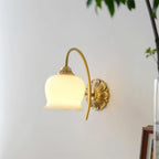 Unlit cream glass flower shade on a curved brass arm, a Clérachic wall light on a pale wall with a plant leaf at the edge.