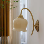 Cream glass flower shade on a brass arm, a Clérachic wall light beside gold curtains and a window.