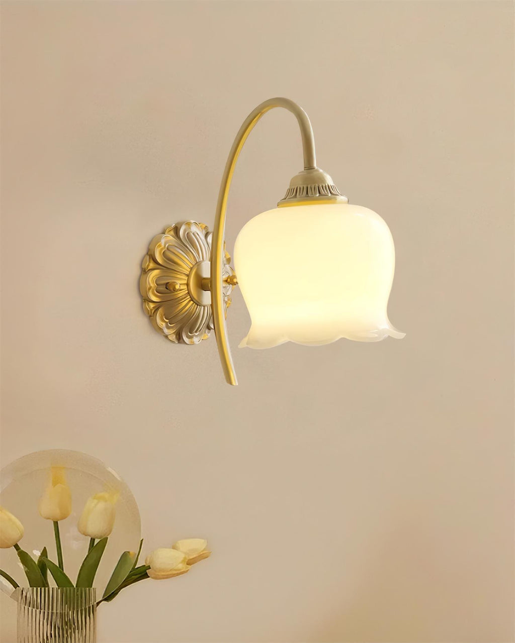 Lit Clérachic wall light with a flower-shaped glass shade on a peach wall, above a vase of yellow tulips.