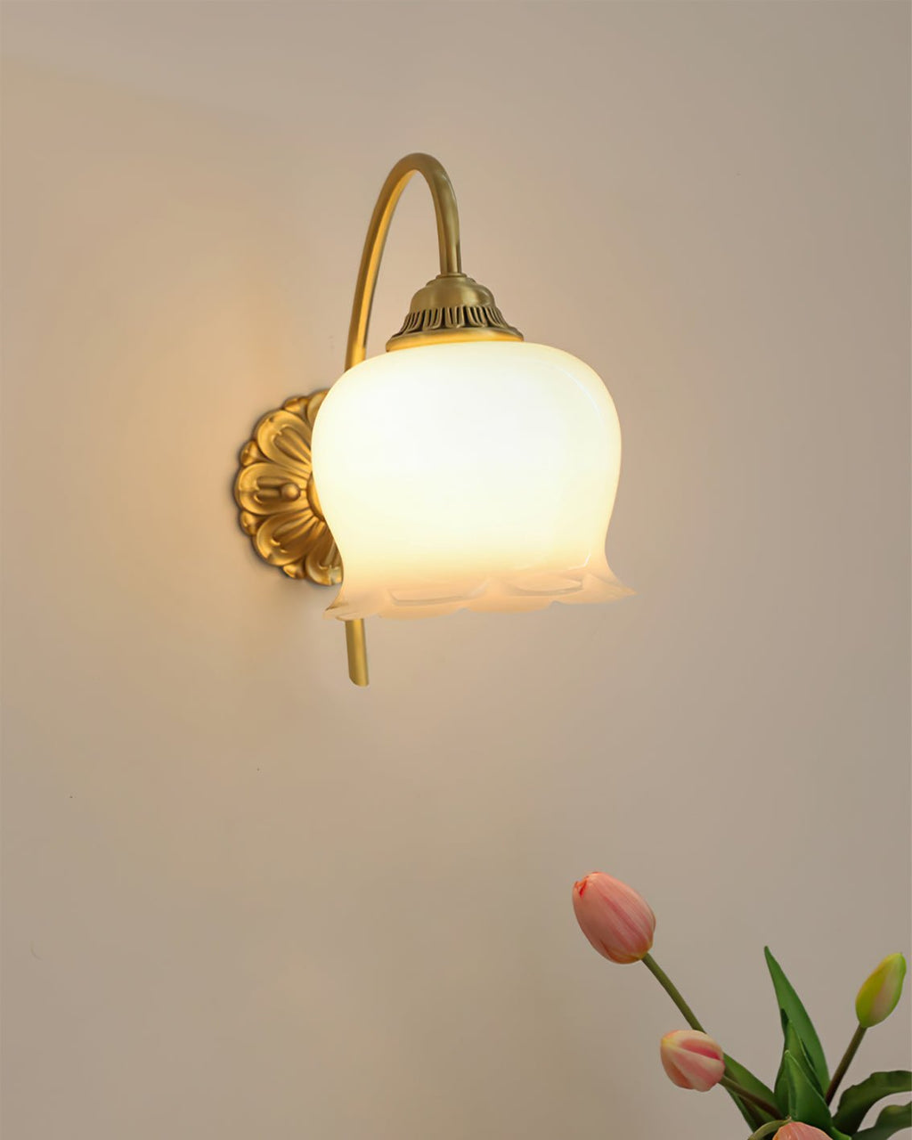 Clérachic wall light lit warm on a plain wall, with the tip of a pink tulip at the corner of the frame.