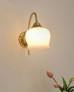 Clérachic wall light lit warm on a plain wall, with the tip of a pink tulip at the corner of the frame.