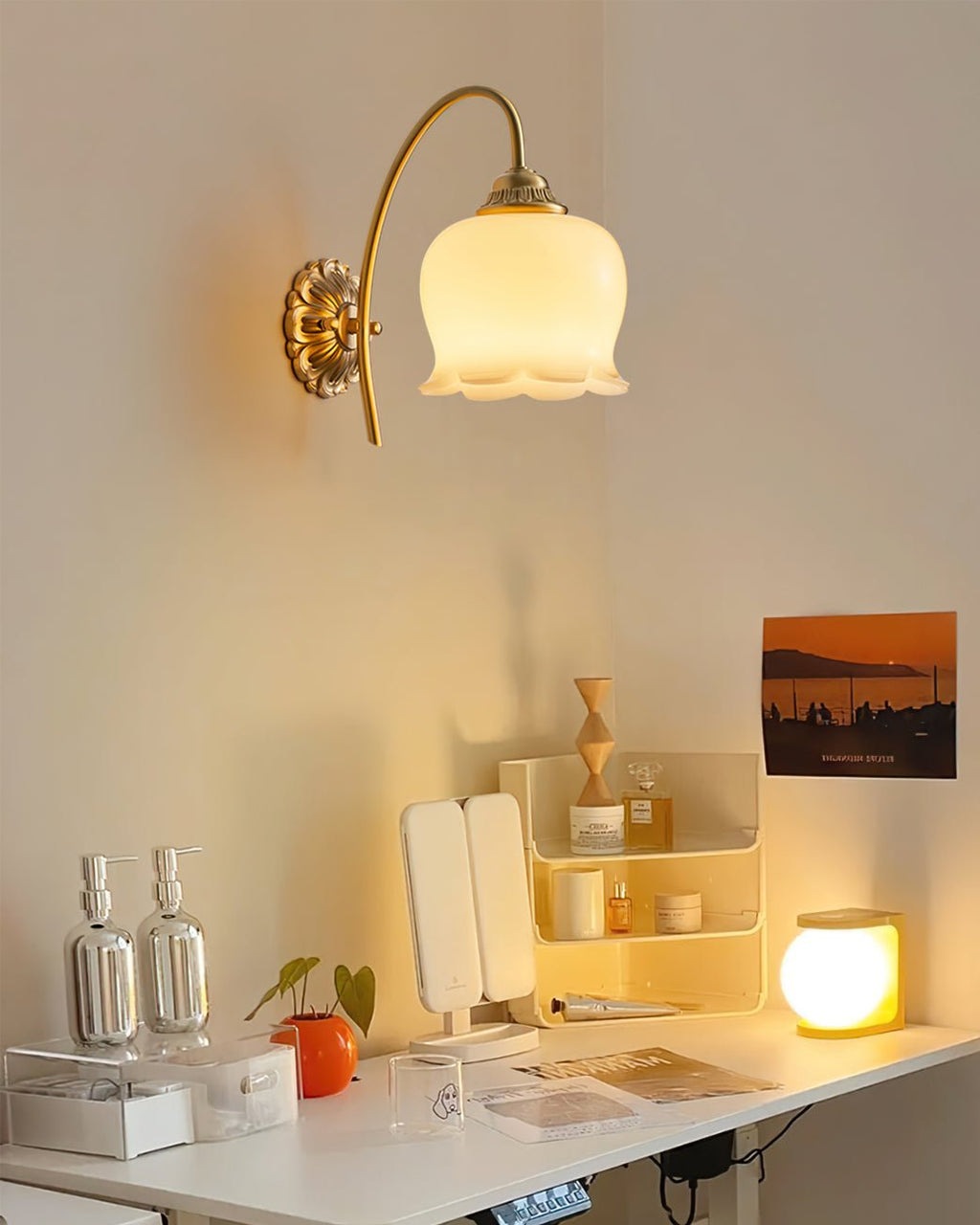Clérachic brass wall light lit above a desk with a vanity mirror, a small lamp and a sunset poster.