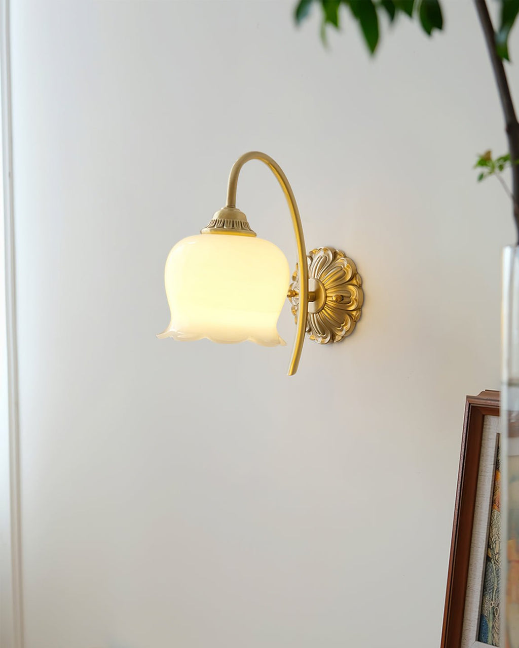 Lit Clérachic wall light on a white wall, a leafy plant and the edge of a picture frame nearby.