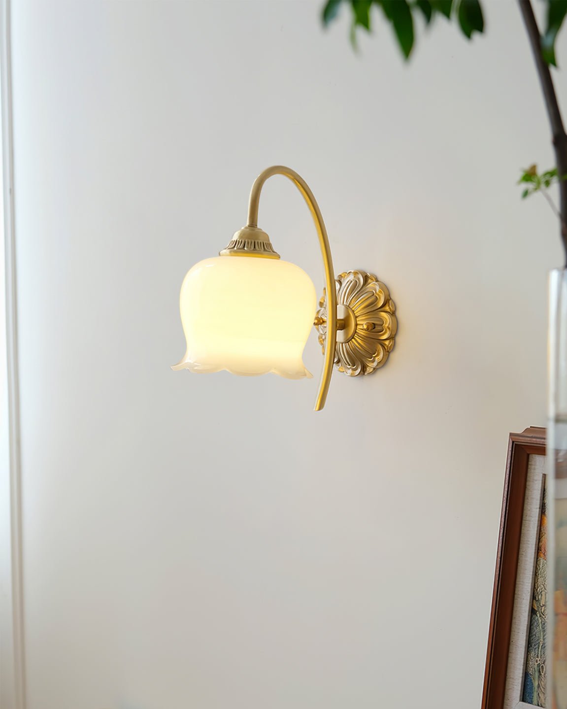 Lit Clérachic wall light on a white wall, a leafy plant and the edge of a picture frame nearby.