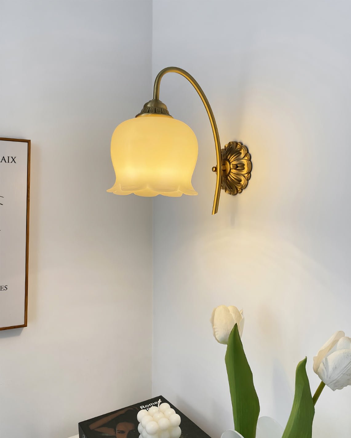 Clérachic wall light lit in a white room corner, above a vase of white tulips and a framed print.