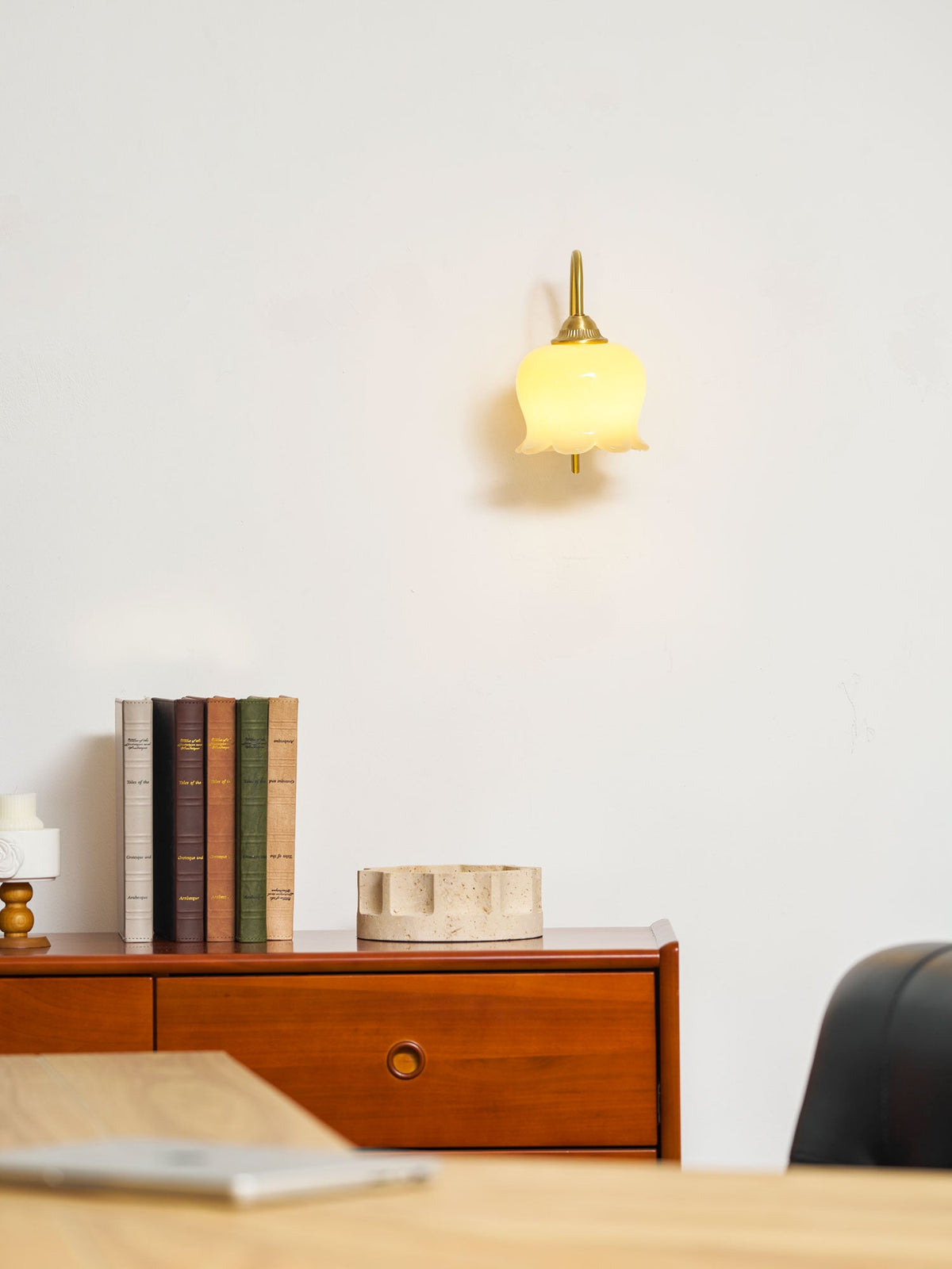 Small lit Clérachic wall light high on a white wall, above a wooden chest of drawers with books and a candle.