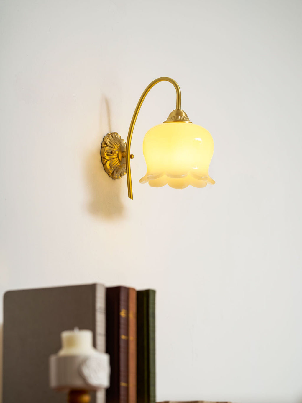 Close, lit view of the Clérachic wall light with its scalloped glass shade, above a blurred stack of books.