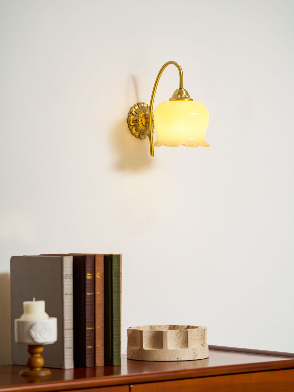 Clérachic wall light lit warm on a white wall above a stack of books, a candle and a stone tray.