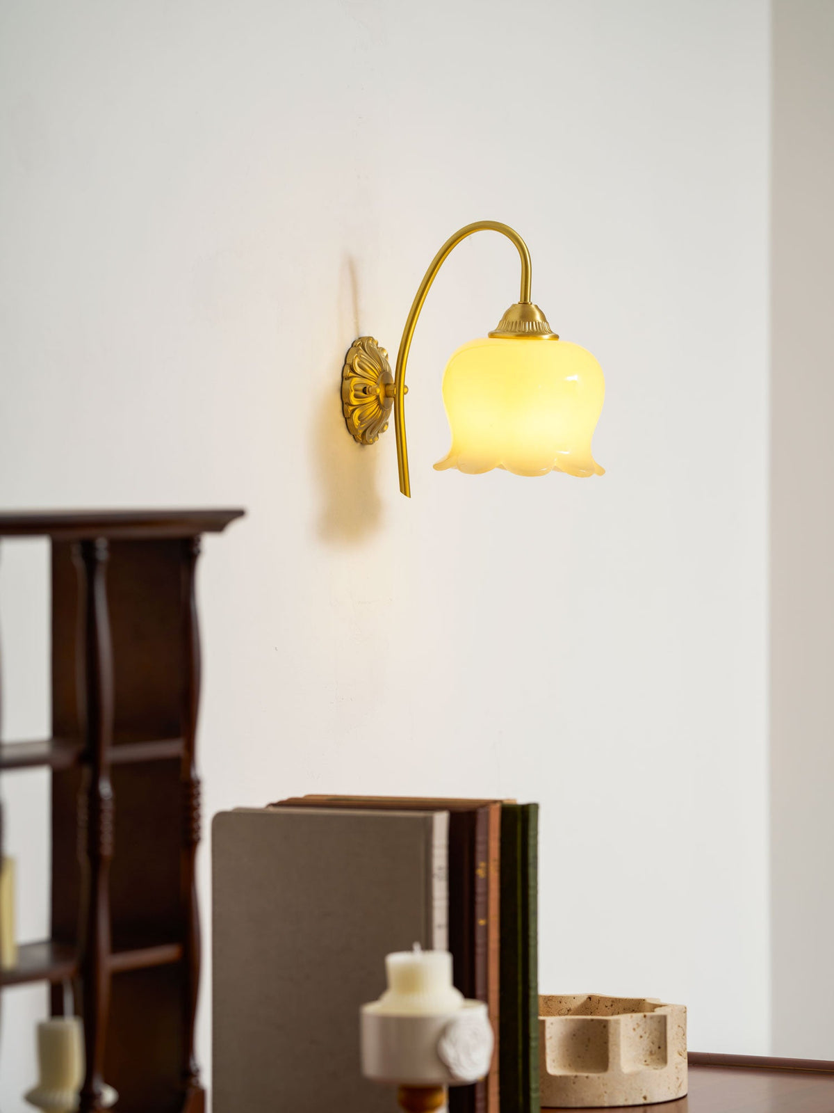 Clérachic wall light lit warm above a dark wooden shelf unit and a stack of books on a white wall.