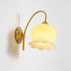 Lit Clérachic wall light with a scalloped glass shade, glowing yellow-white, against a plain white wall.