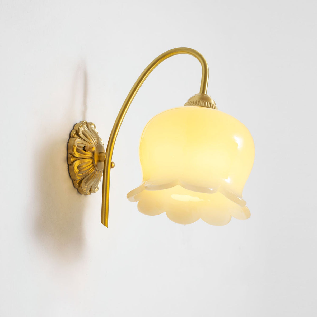 Lit Clérachic wall light with a scalloped glass shade, glowing yellow-white, against a plain white wall.