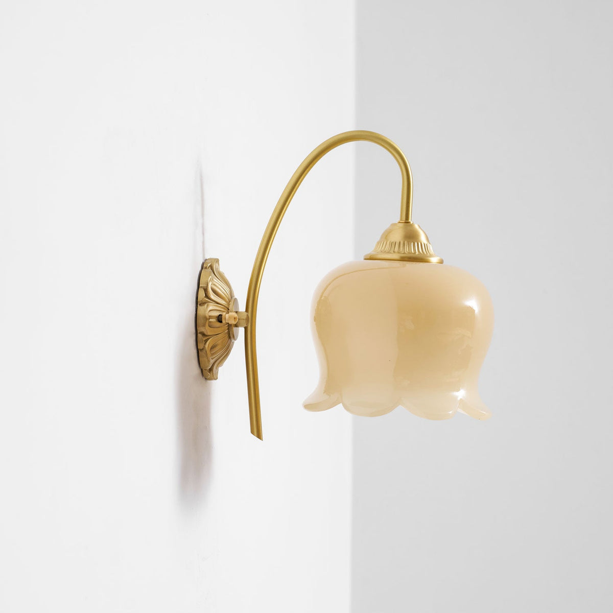 Unlit Clérachic wall light with a cream glass shade on a curved brass arm, near a wall corner.