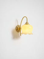 Lit Clérachic wall light with a soft glow, its brass arm casting a shadow on a pale grey wall.