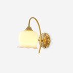 Clérachic brass wall light lit on a pale background, with a bell-shaped cream glass shade and ornate back plate.