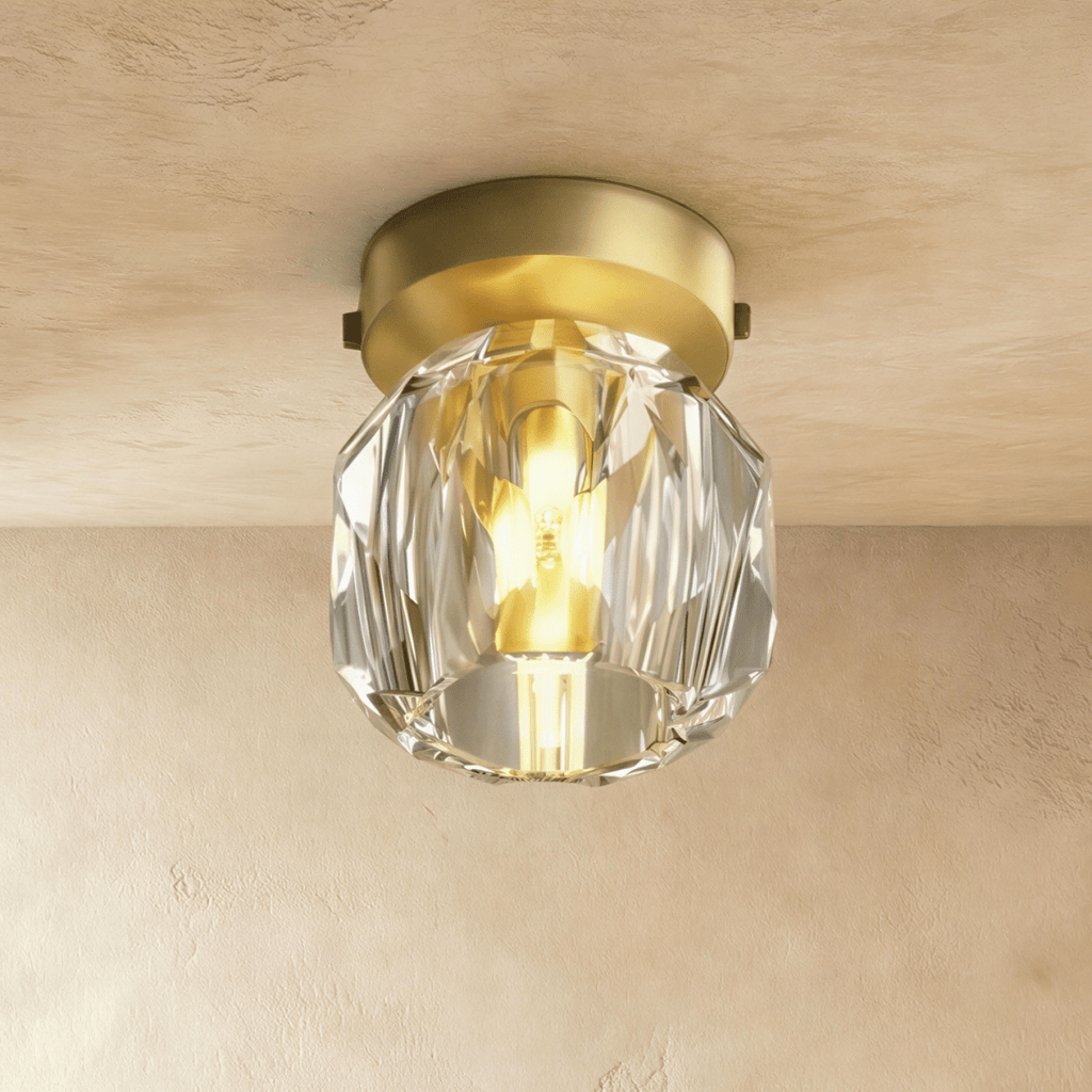 Clérachic ceiling light in close-up, a faceted crystal shade glowing around its bulb beneath a gold ceiling plate.