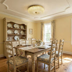 Glowing Clérachic glass ceiling light in a yellow dining room with a plate dresser, a wooden table and ladder-back chairs.