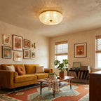 Clérachic flush ceiling light lit above a warm living room with a mustard sofa, a rust and teal rug and framed prints on the wall.