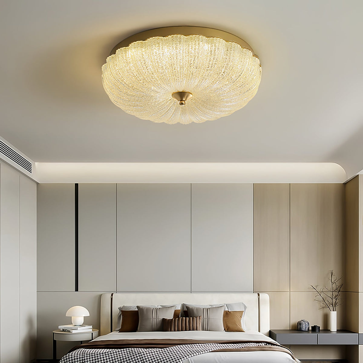 Lit Clérachic flush ceiling light above a bedroom with a pale headboard, brown cushions and a small lamp on a dark bedside table.