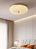 Clérachic ceiling light glowing near a cove-lit wall, with a bed with orange cushions and a round table lamp below.