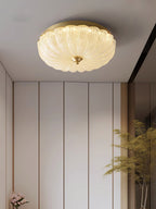 Clérachic flush ceiling light lit above cream wall panels, with a tall plant and pink flowers at the edge of the frame.