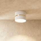 Clérachic white surface-mounted downlight with a slim brass ring accent, glowing on a plaster ceiling.