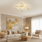 Gold four-loop Clérachic ceiling light lit above a cream living room, with a beige sofa, a round wooden coffee table and sheer curtains.