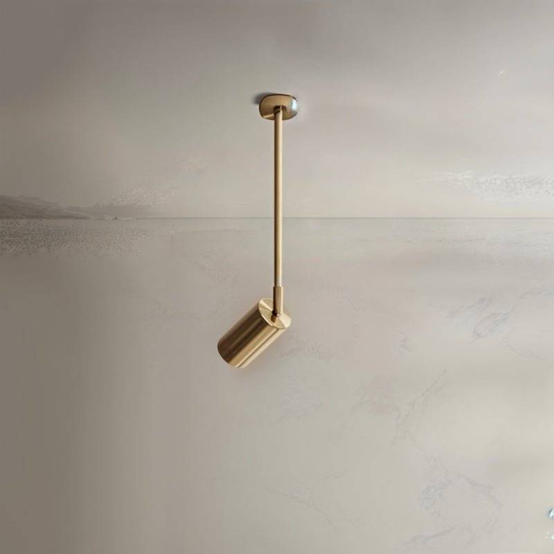 Clérachic gold ceiling spotlight on a longer rod with a small cylinder head, aimed down against a pale plaster wall.