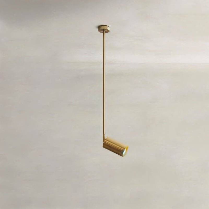 Clérachic gold ceiling spotlight hanging from a slim gold rod, its small cylinder head angled and lit on a plaster backdrop.
