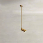 Clérachic gold ceiling spotlight hanging from a slim gold rod, its small cylinder head angled and lit on a plaster backdrop.