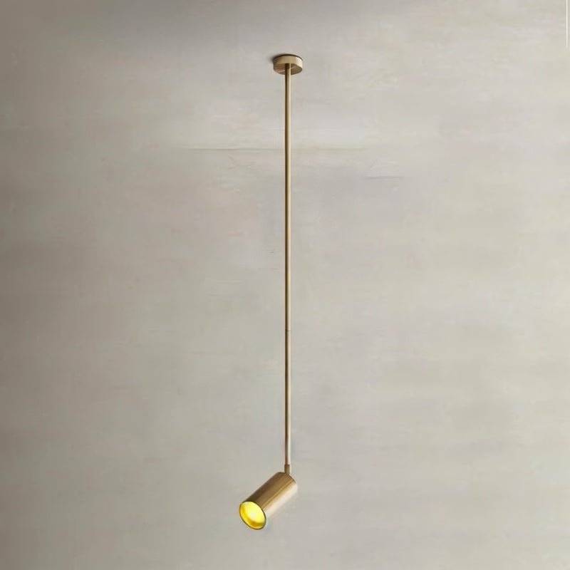 Clérachic gold ceiling spotlight on a very long thin gold rod, its cylinder head tilted and glowing against a stone-toned wall.