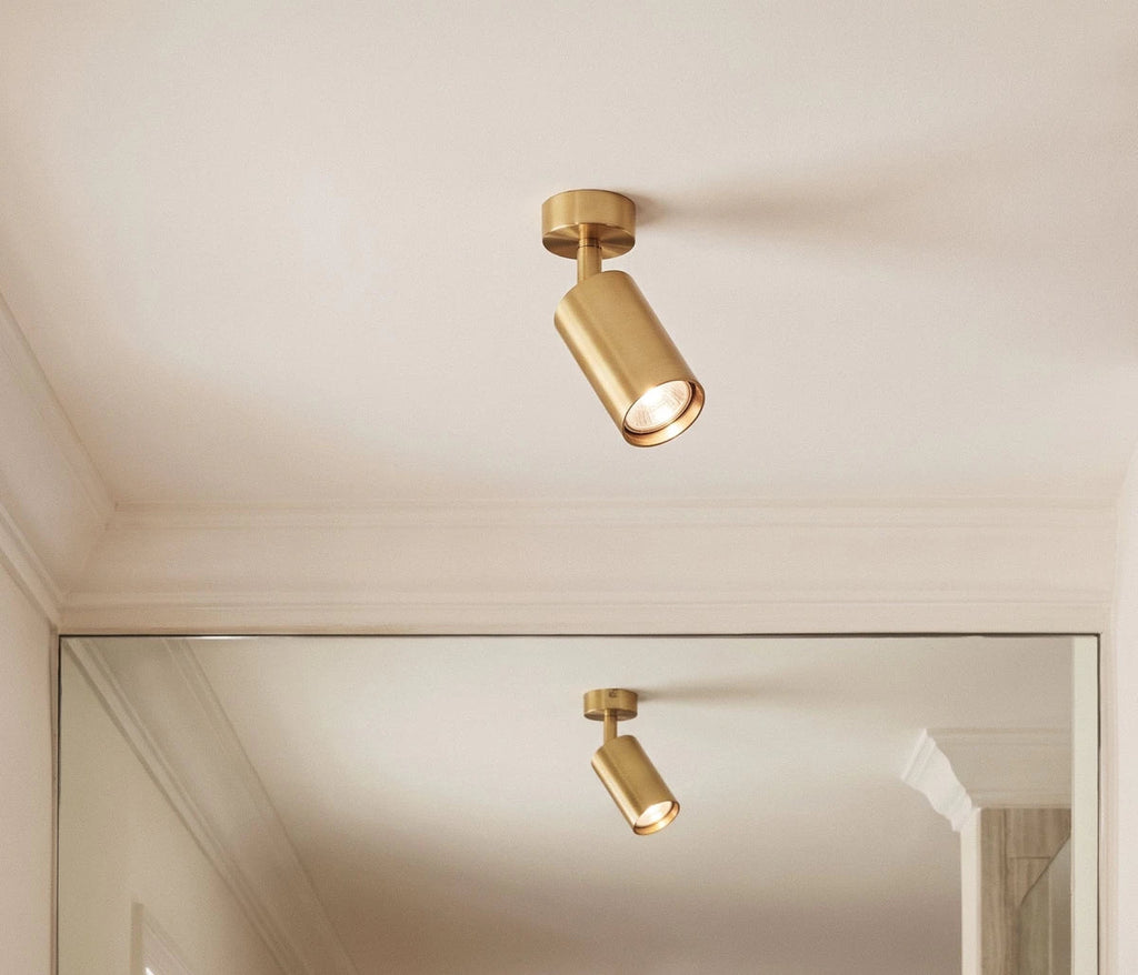 Clérachic gold ceiling spotlight with a tilting cylinder head lit on a white ceiling, reflected in a mirror below.