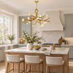 A lit Clérachic gold branch chandelier above a marble kitchen island with linen stools and sage-blue cabinets behind.